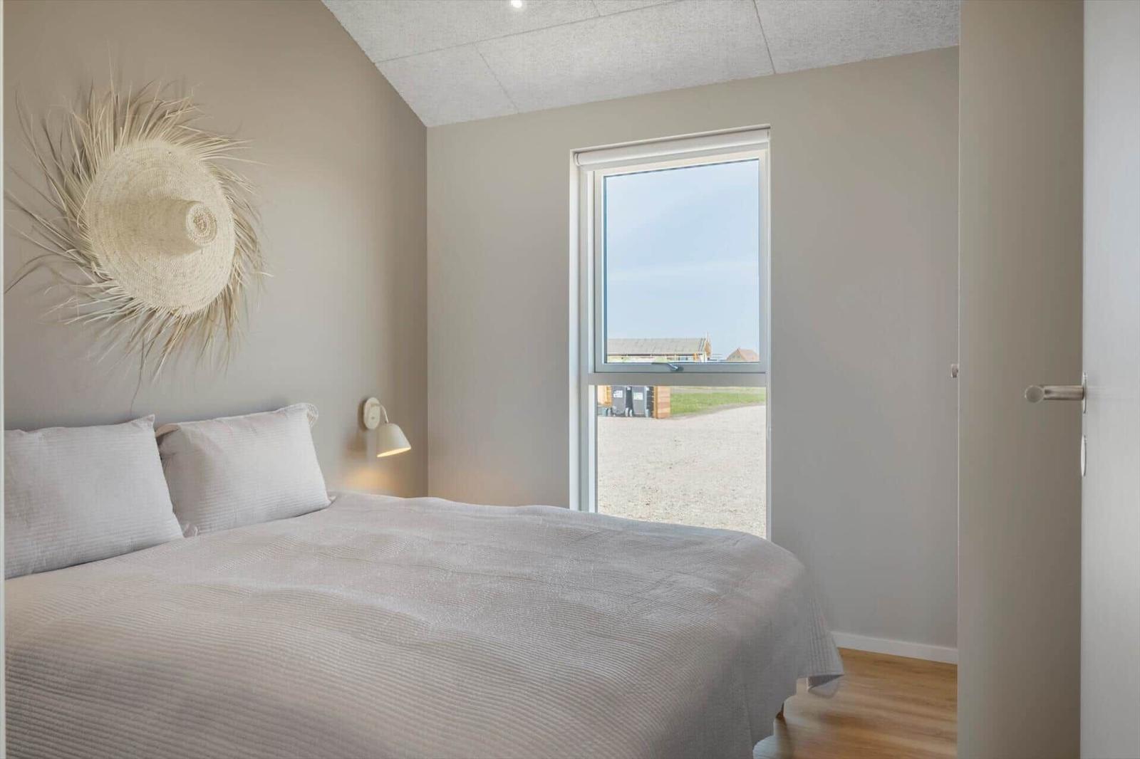 Schlafzimmer mit Bett, Wandlampe und Fensterblick auf Außenbereich.