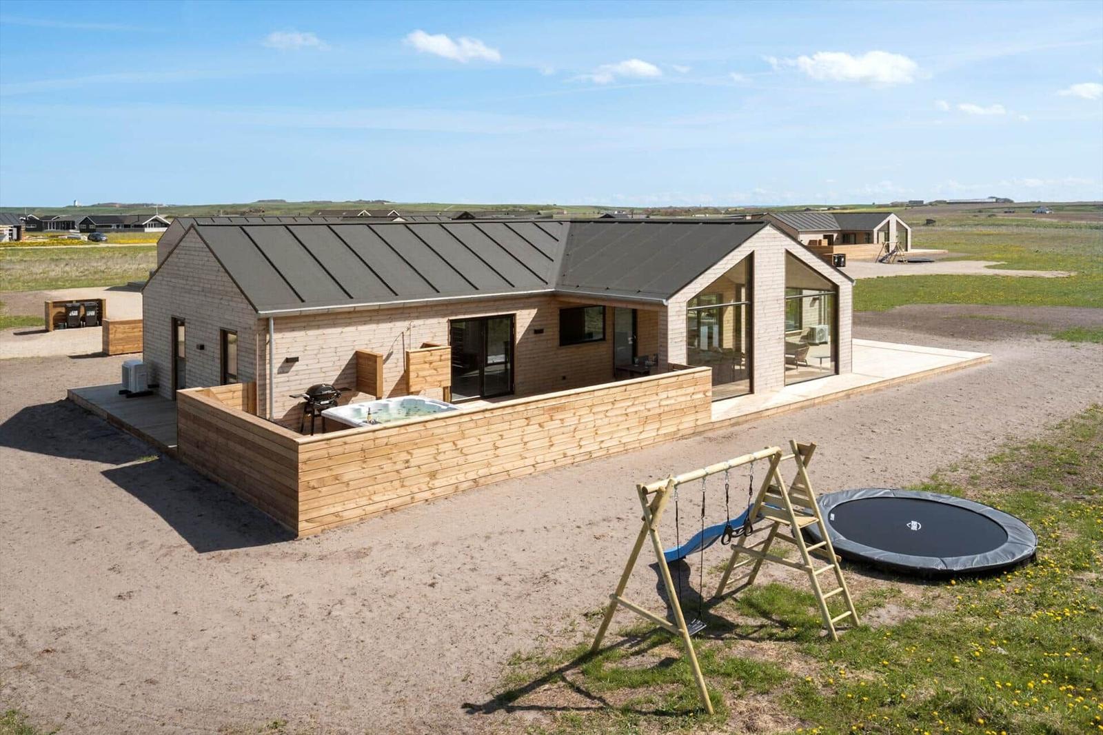 Moderne Holzhütte mit Whirlpool, Trampolin und Spielplatz auf offener Landschaft.