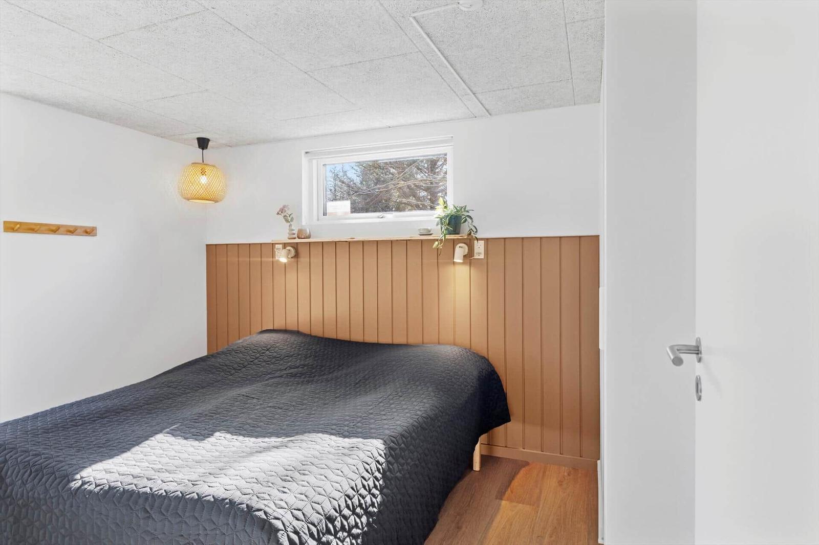 Schlafzimmer mit Bett, Holzwand und Fenster. Decke mit Paneelen, Lampe und Tür sichtbar.