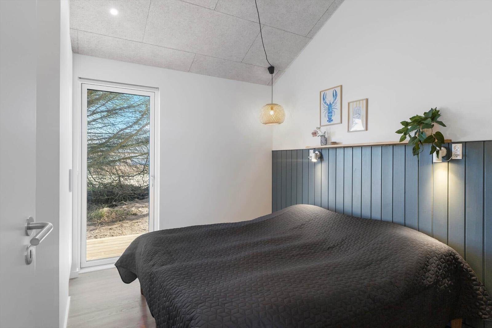 Schlafzimmer mit Bett, Wandverkleidung und Fenster zur Außenterrasse.