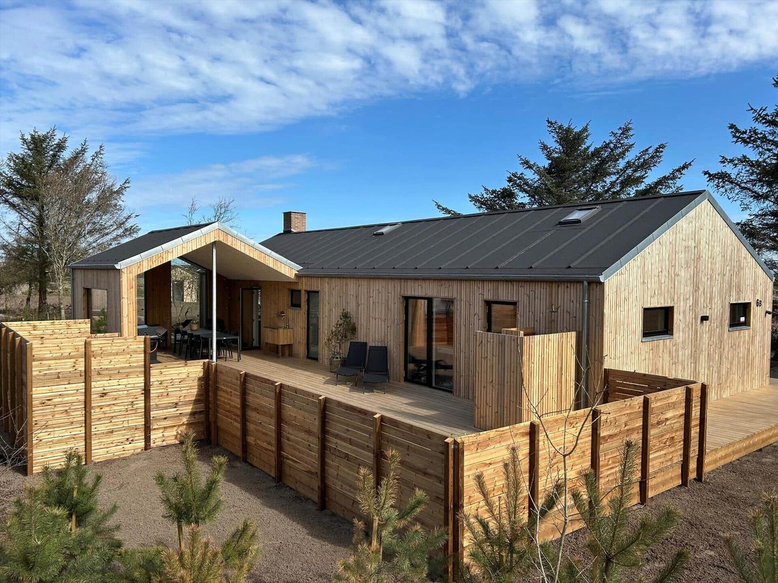 Moderne Holzhütte mit Terrasse und Gartenzaun unter blauem Himmel.