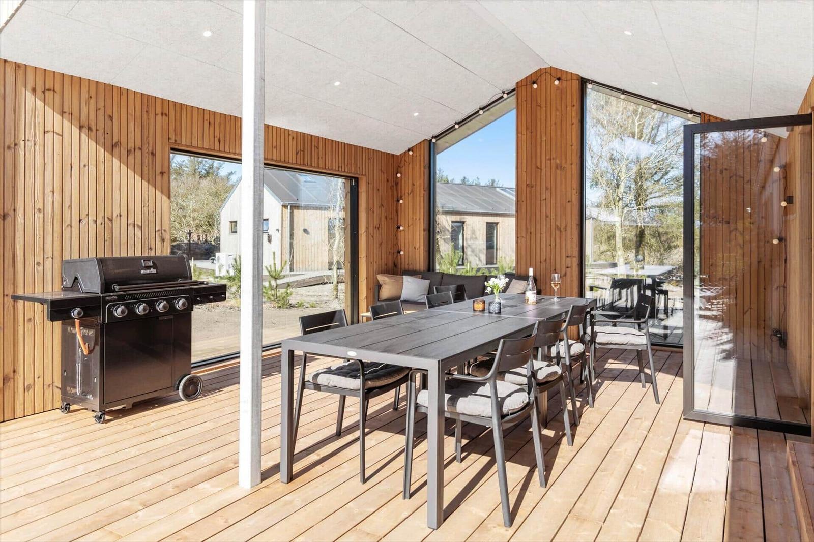 Terrasse mit Esstisch, Grill und Sitzgelegenheiten. Holzwand und große Fenster.