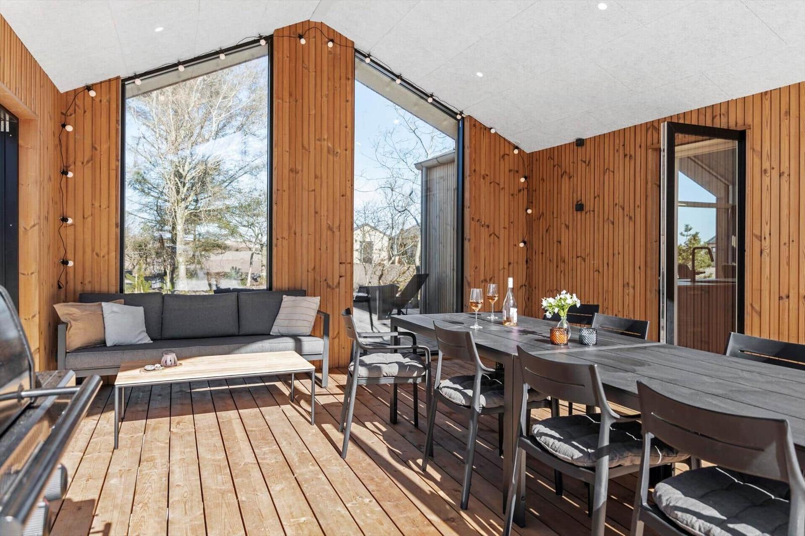 Terrasse mit Sitzgruppe, Esstisch und Grill. Holzwände und große Fenster.