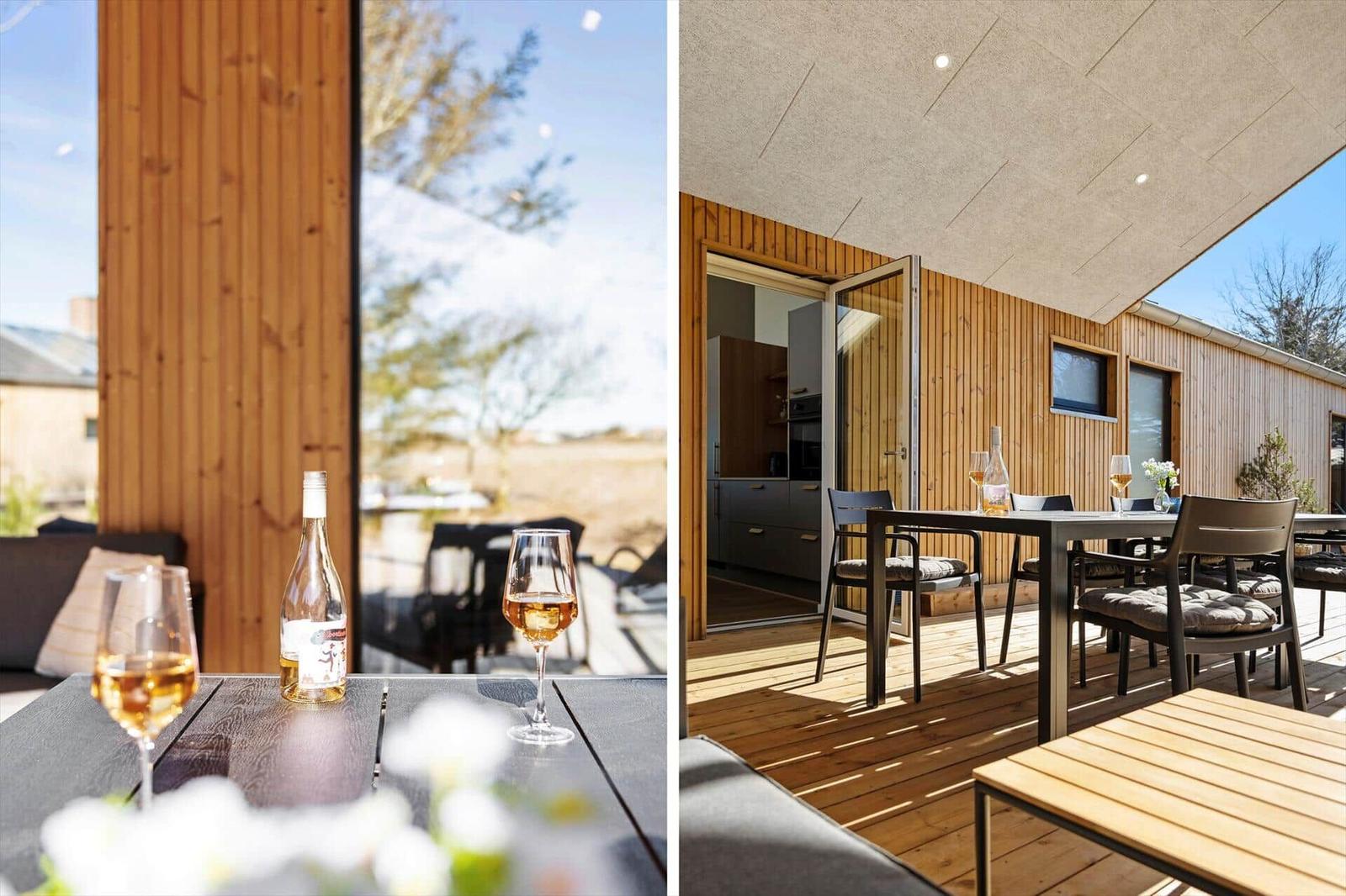 Terrasse mit Tisch, Stühlen und Wein. Holzwand und Blick auf Außengelände.