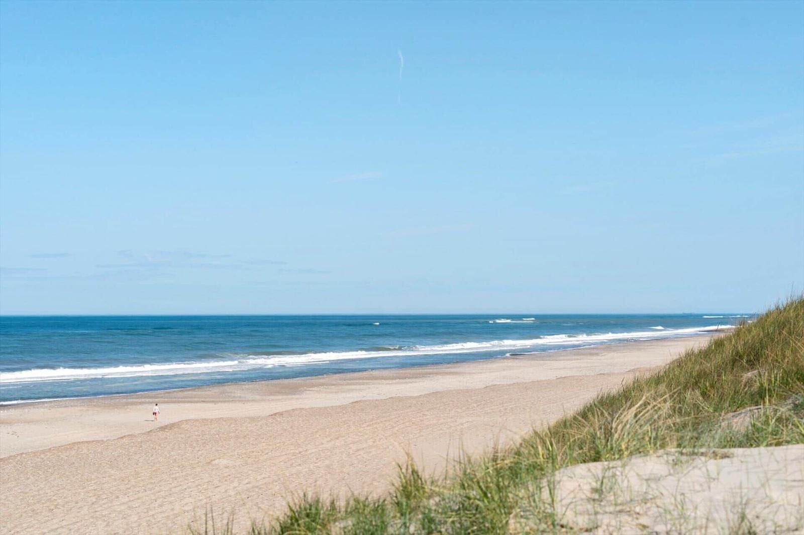 Strand mit Sanddünen und Meer unter blauem Himmel. Ein einzelner Mensch am Ufer.