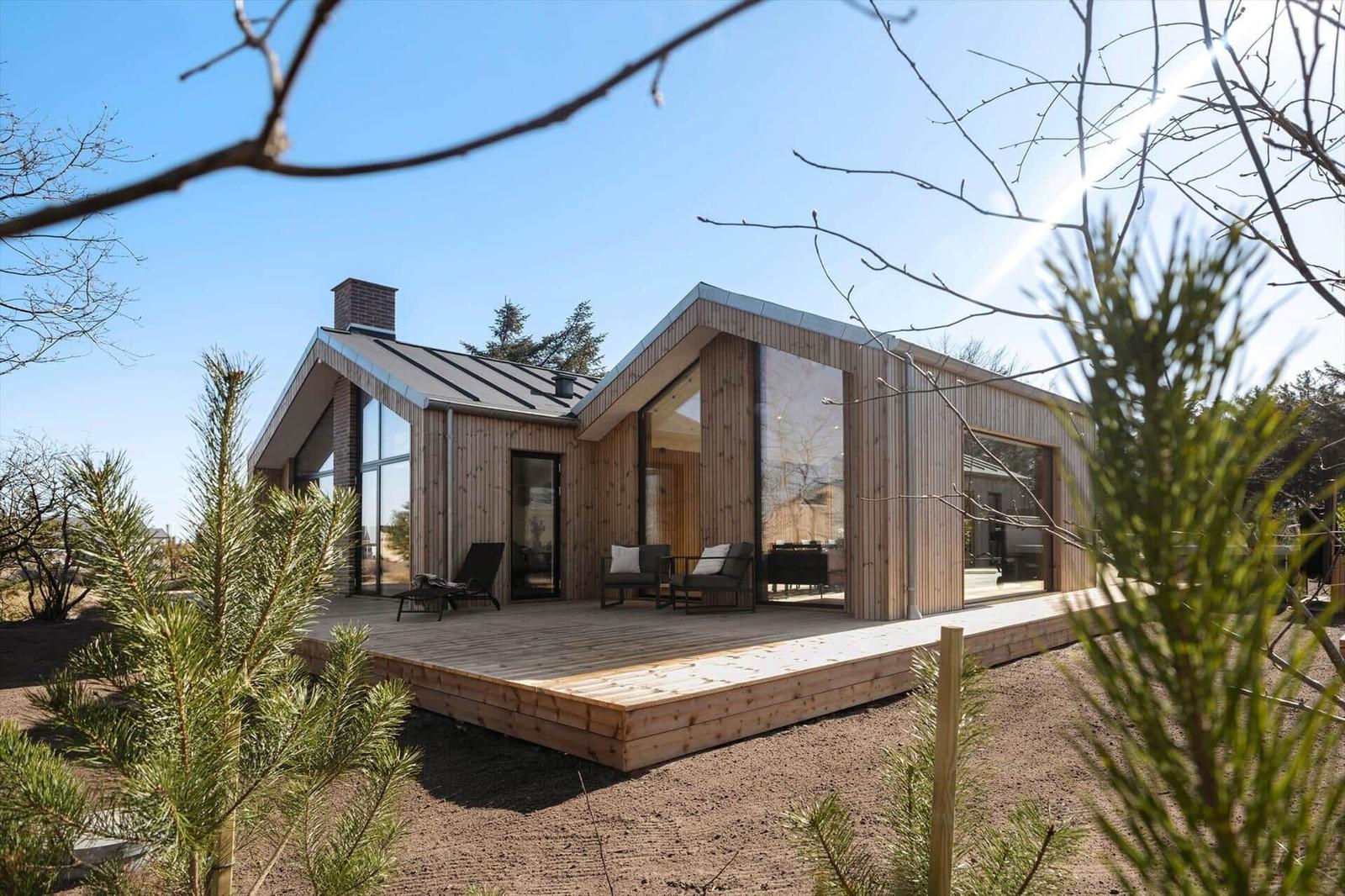 Moderne Holzhütte mit großem Balkon und Glaswänden. Umgeben von Bäumen und klarem Himmel.