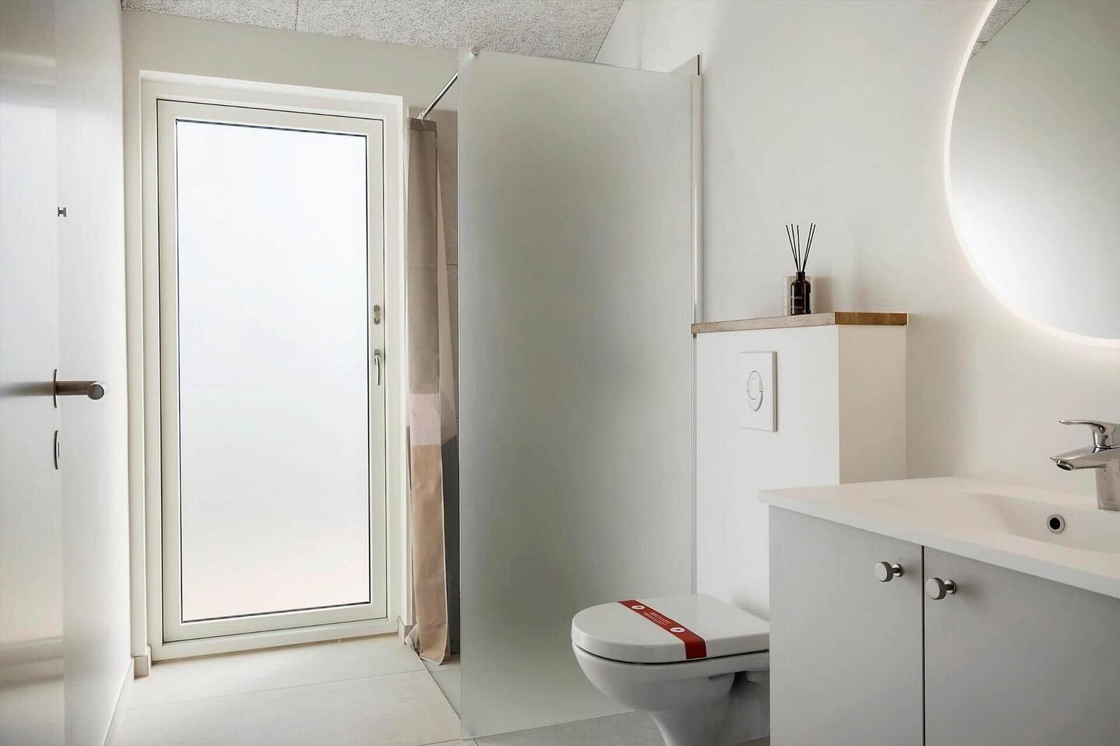 Badezimmer mit Toilette, Waschbecken und Dusche. Fenster mit Milchglas und runder Spiegel.