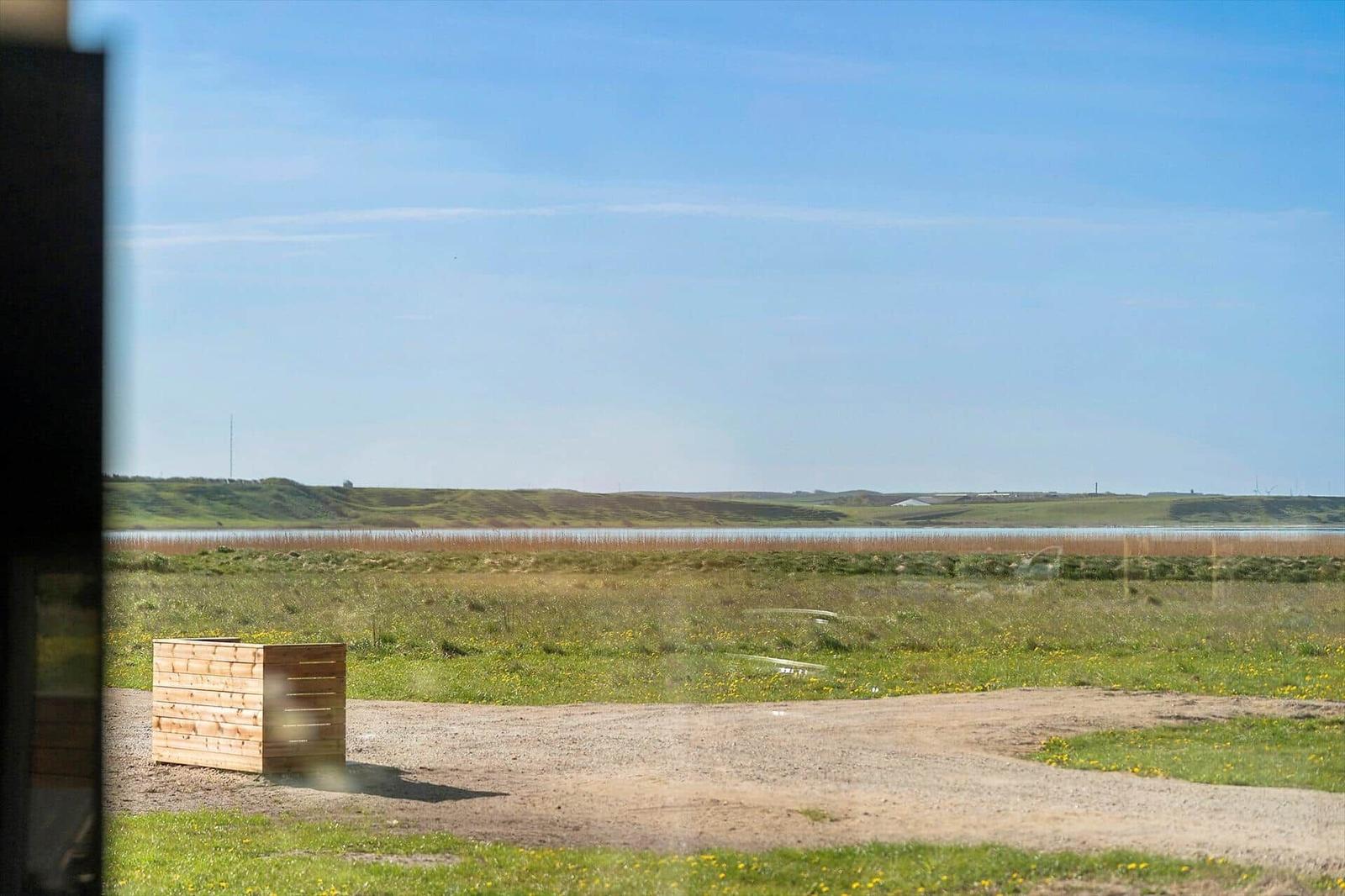 Draußen liegt ein Holzobjekt neben einem unbefestigten Weg. Im Hintergrund erstreckt sich ein See mit Grasland.