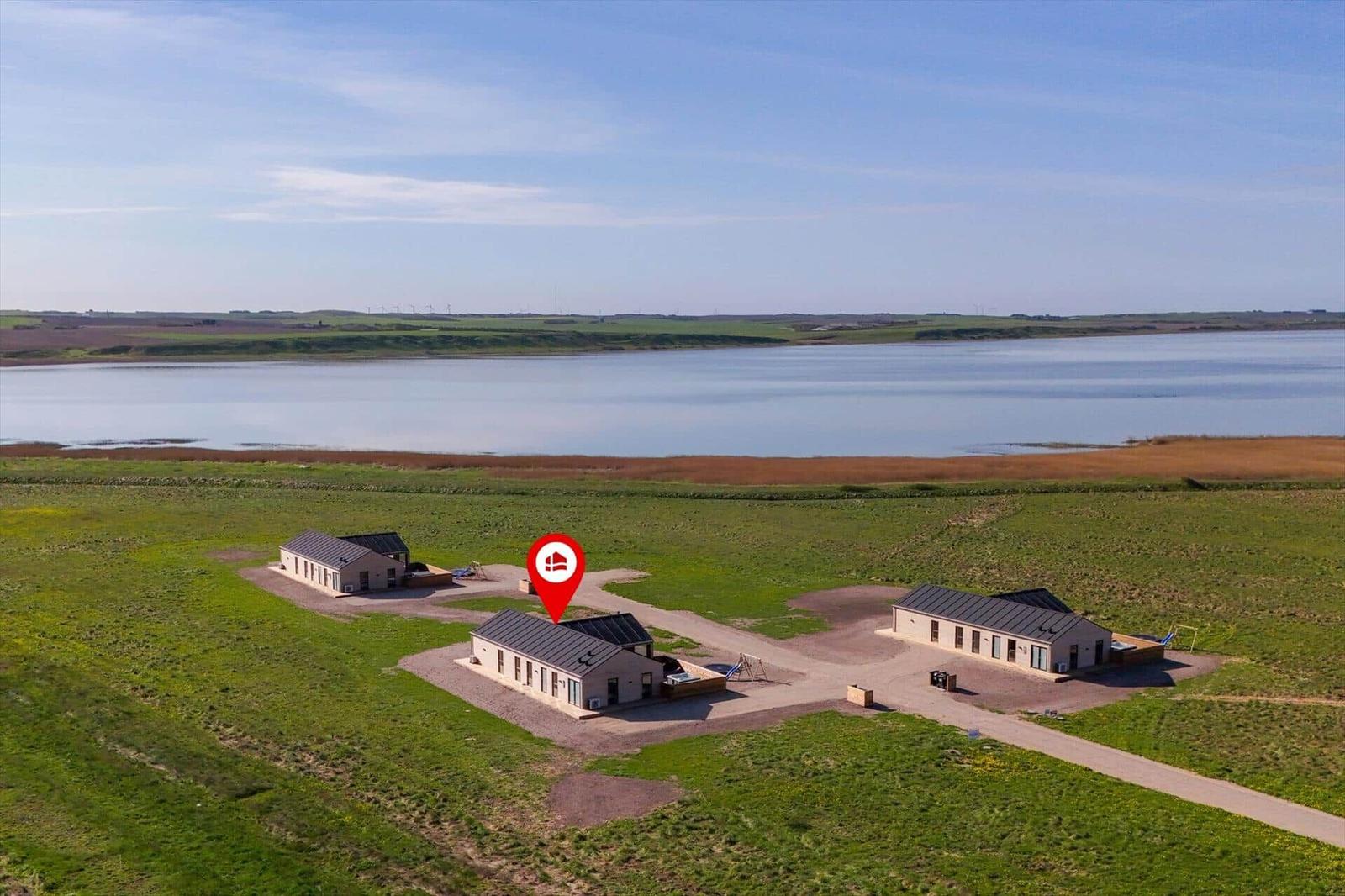 Drei Häuser mit Dach und Terrasse stehen auf einer grünen Wiese neben einem See.