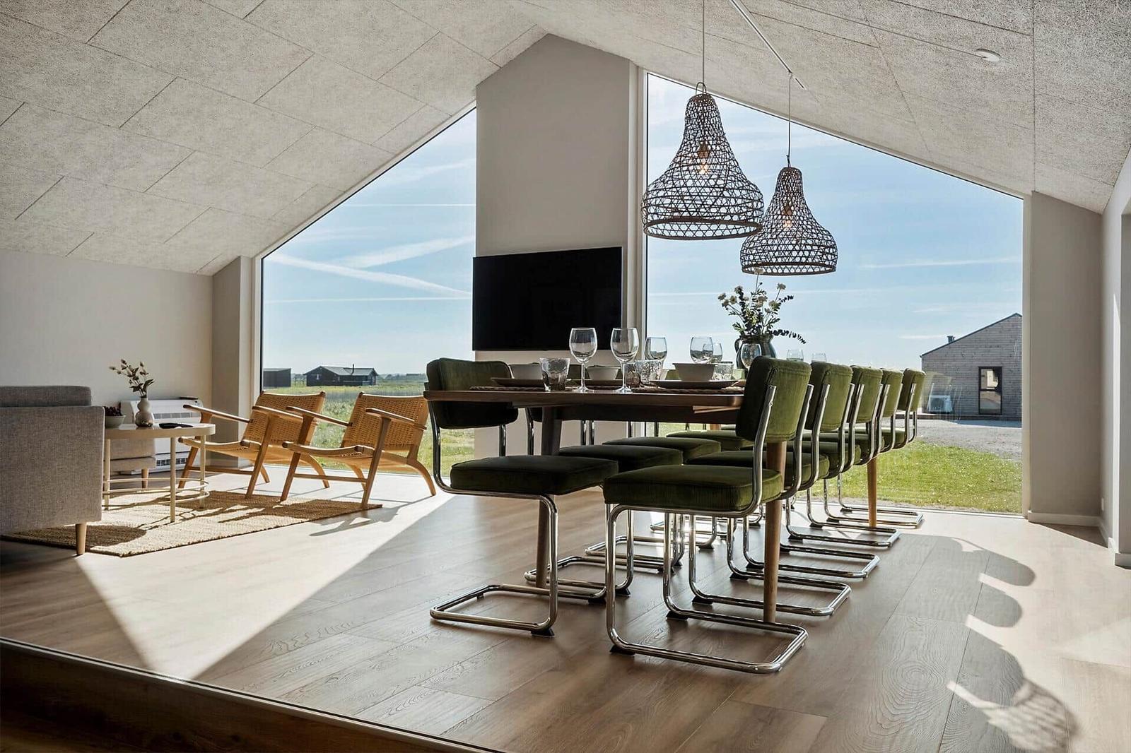 Essbereich mit langem Tisch und grünen Stühlen unter großen Fenstern mit Blick auf die Landschaft.