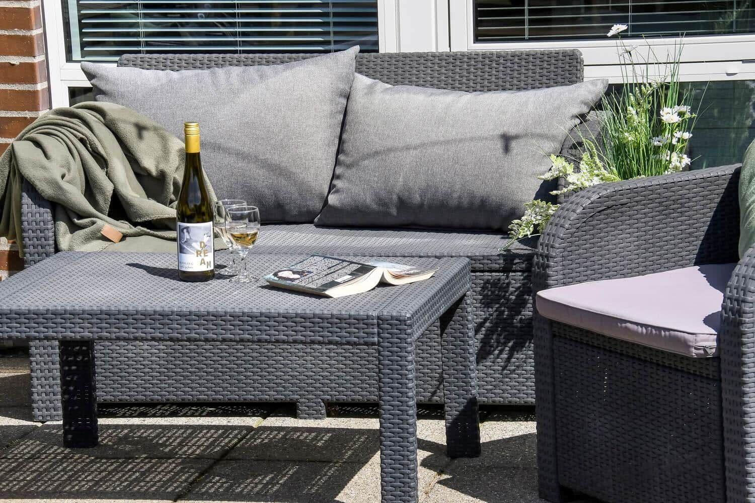 Außenbereich mit grauem Rattansofa, Tisch, Flasche und Buch.