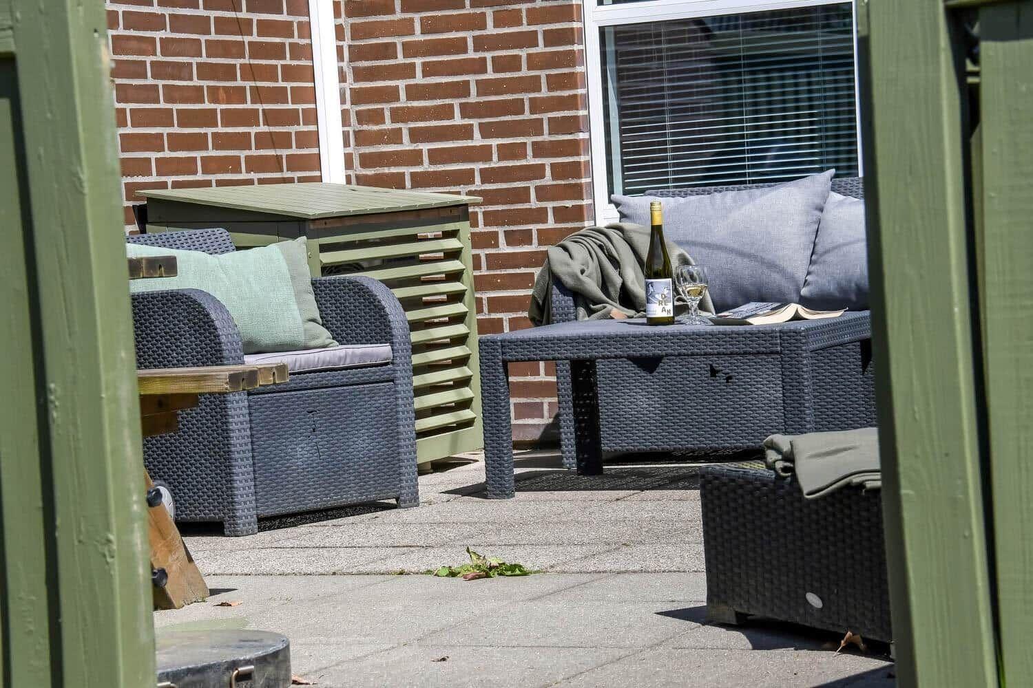 Terrasse mit Gartenmöbeln, Tisch und Weinflasche vor Backsteinmauer.