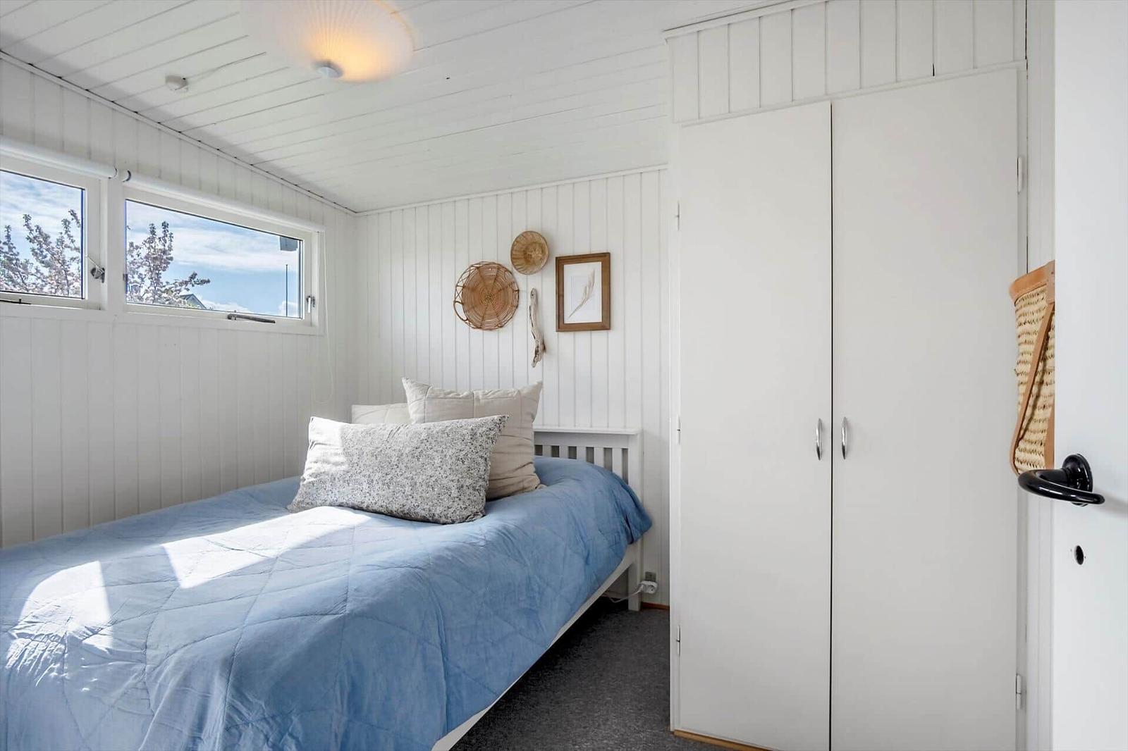 Schlafzimmer mit Bett, Kleiderschrank und Fenster. Wände und Decke weiß verkleidet.