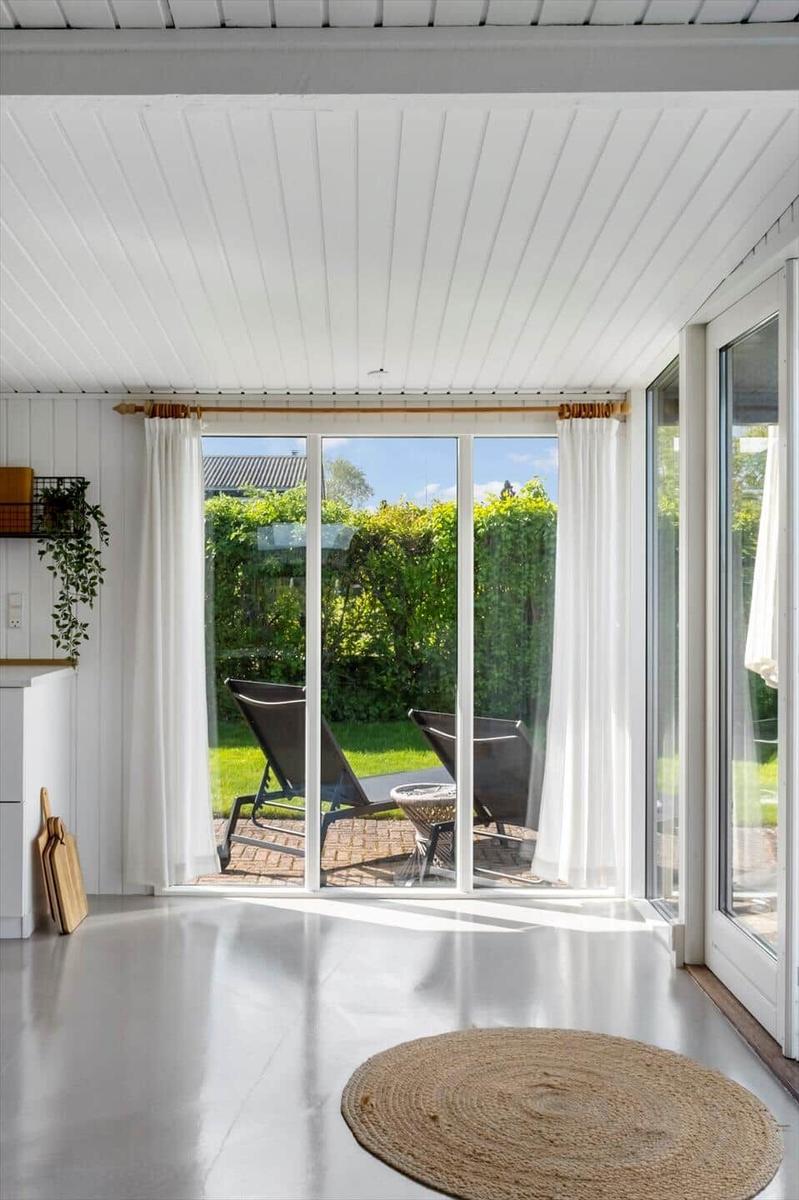 Große Fensterfront mit Blick auf Garten und Liegestühle. Weiße Wände und Boden. Runder Teppich.
