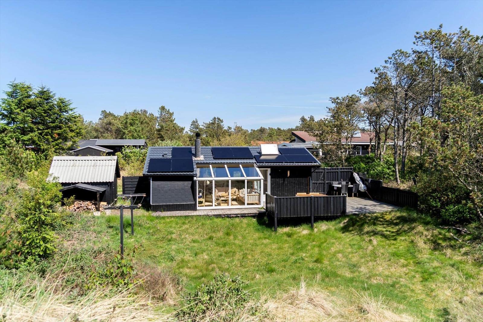 Schwarze Holzhütte mit Glasveranda und Solarpanel auf Dach. Umgeben von Gras und Bäumen.
