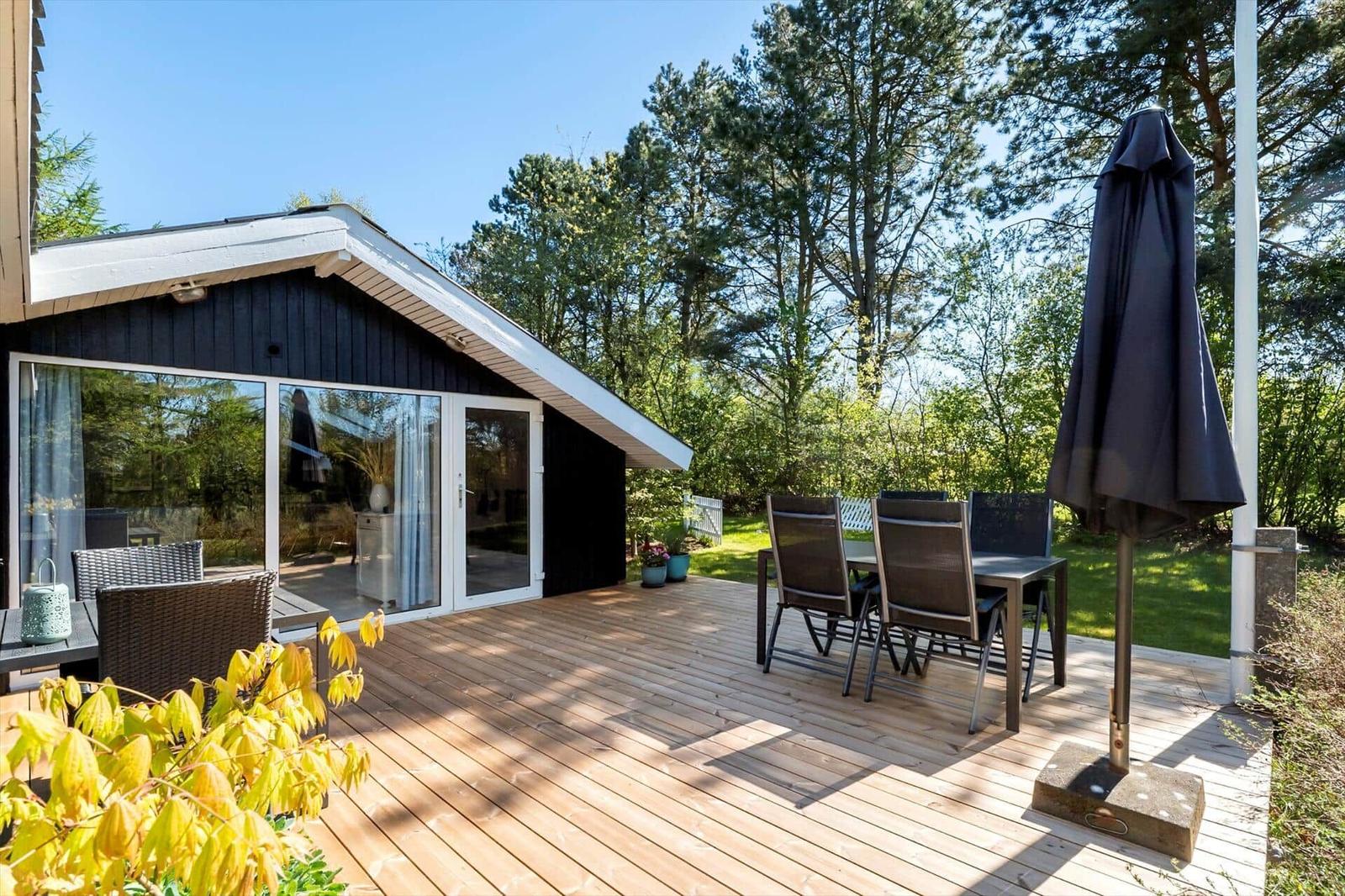 Deck with table, chairs, and closed umbrella. Background: forest and house with glass facade.