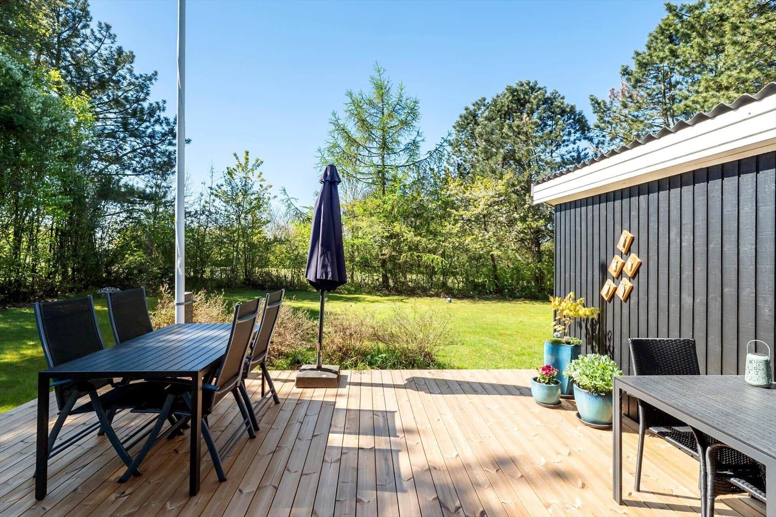 Wooden terrace with table, chairs, and umbrella. Green areas and trees in the background.