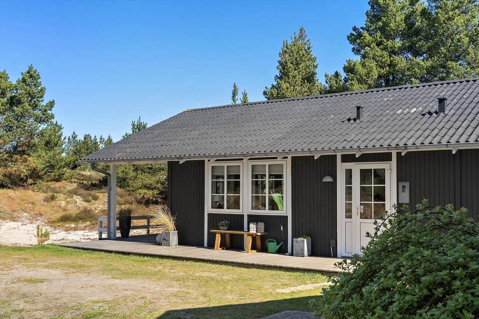 Ein Haus mit dunkler Fassade und weißer Tür. Auf der Terrasse steht ein Holztisch. Umgeben von Bäumen und Gras.