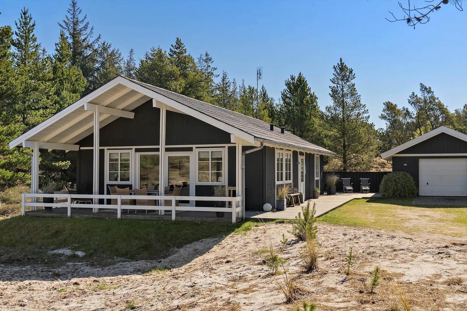 Moderne Holzhütte mit Terrasse, Garten und Garage. Umgeben von Nadelbäumen und Sand.