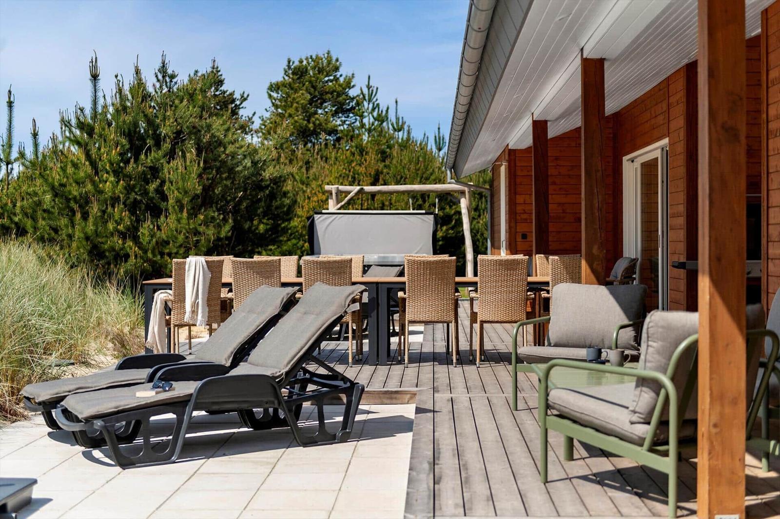 Terrasse mit Liegestühlen, Tisch und Stühlen, Holzwand und Grill unter Bäumen.