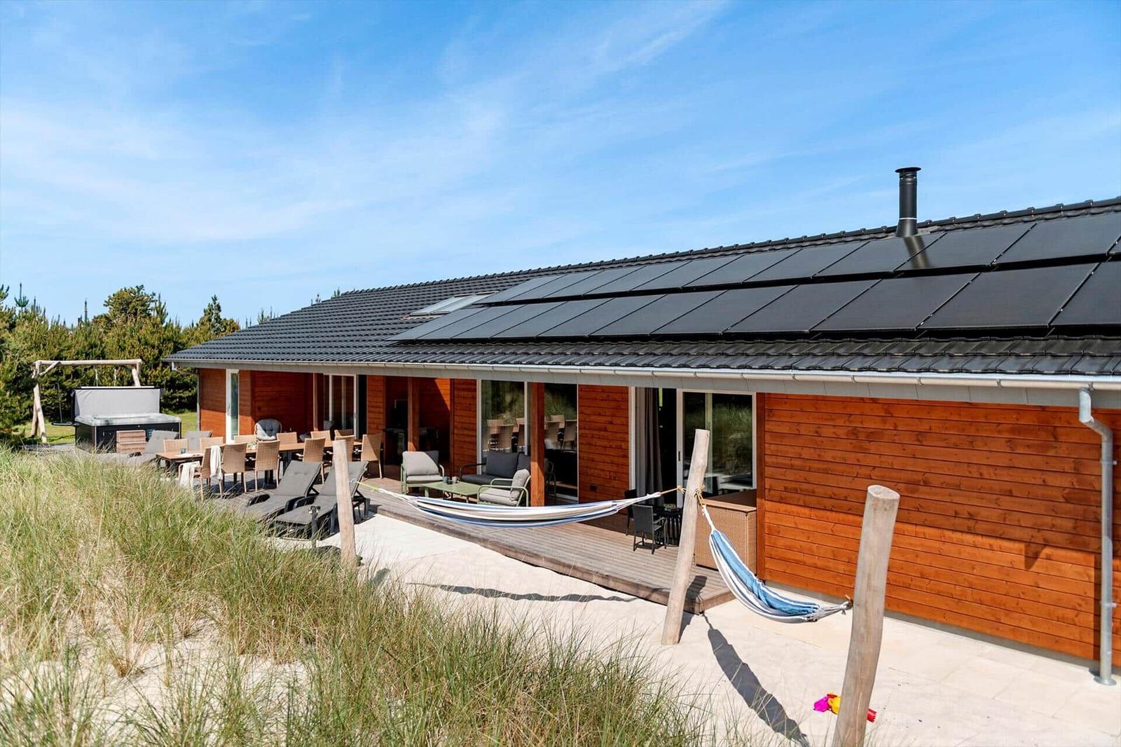 Holzhaus mit Terrasse, Liegen und Hängematten. Dach mit Solarpanele. Umgeben von Gras und Bäumen.