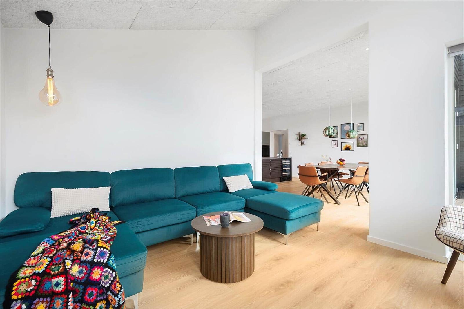 Ein Wohnzimmer mit türkisfarbenem Sofa, Holzboden und runden Couchtisch. Im Hintergrund ist ein Essbereich.