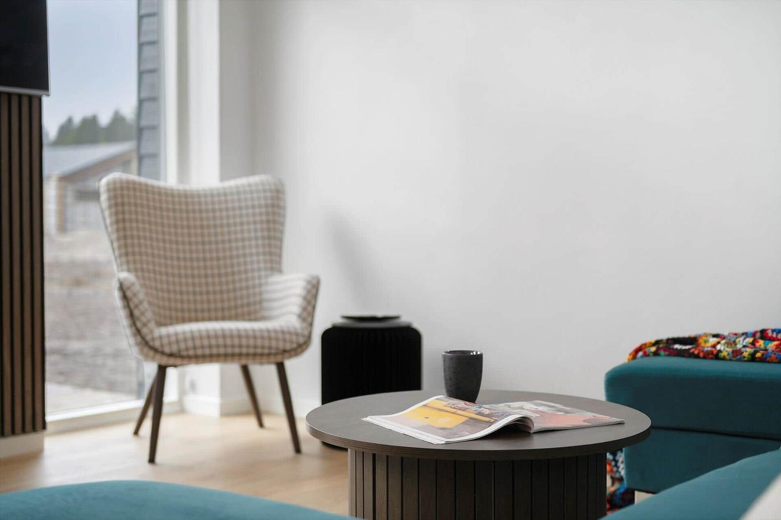 Rundes Tisch mit Zeitschrift und Tasse. Hocker, Sessel und Fenster sichtbar.