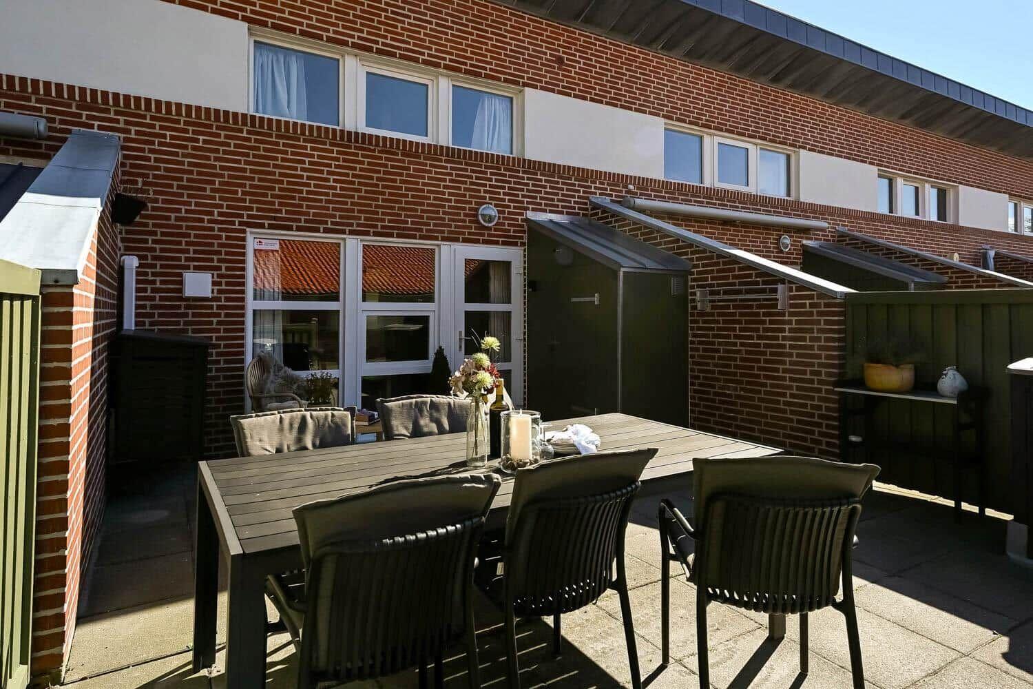 Terrasse mit Tisch und Stühlen vor einem Backsteinhaus mit Fenstern und Sonnenschutz.