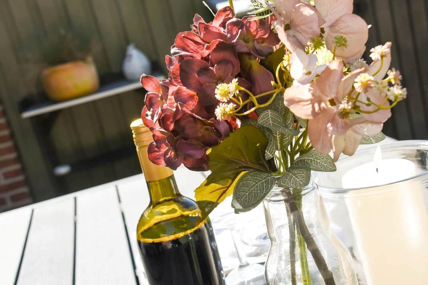 Auf dem Tisch steht eine Blumenvase mit roten und rosa Hortensien, eine Weinflasche und eine Kerze.
