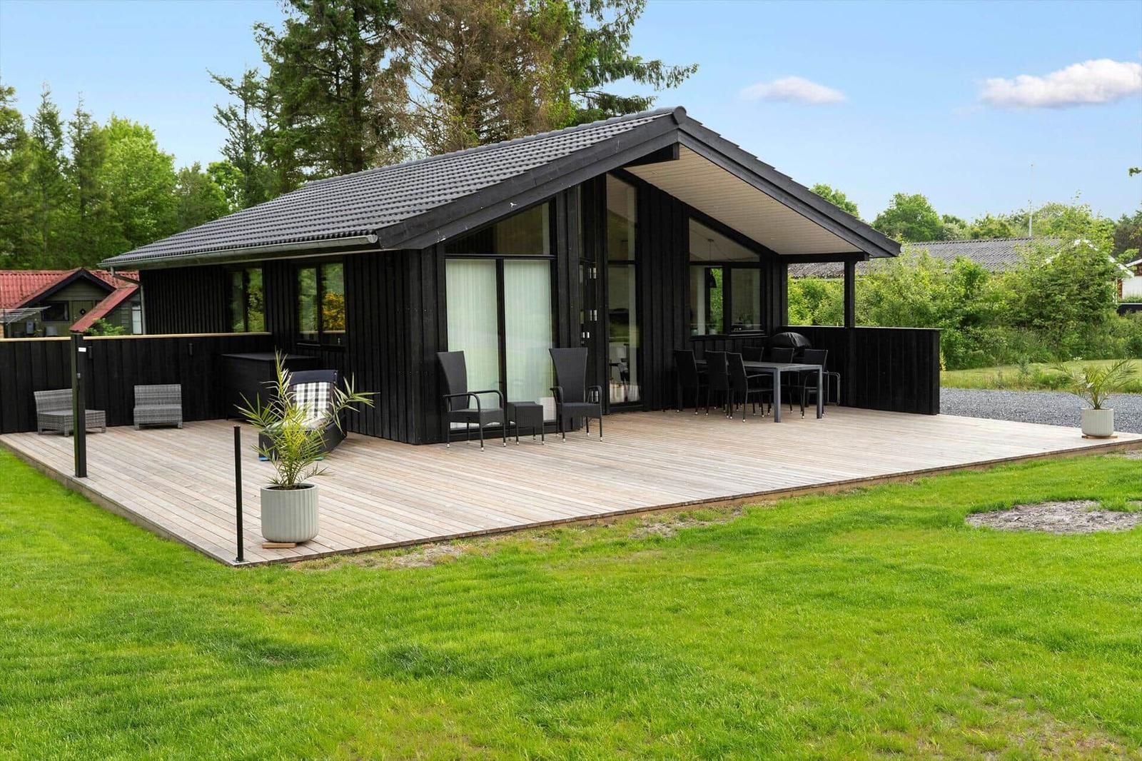 Modern wooden cabin with large terrace and garden. Garden furniture and plants are present.