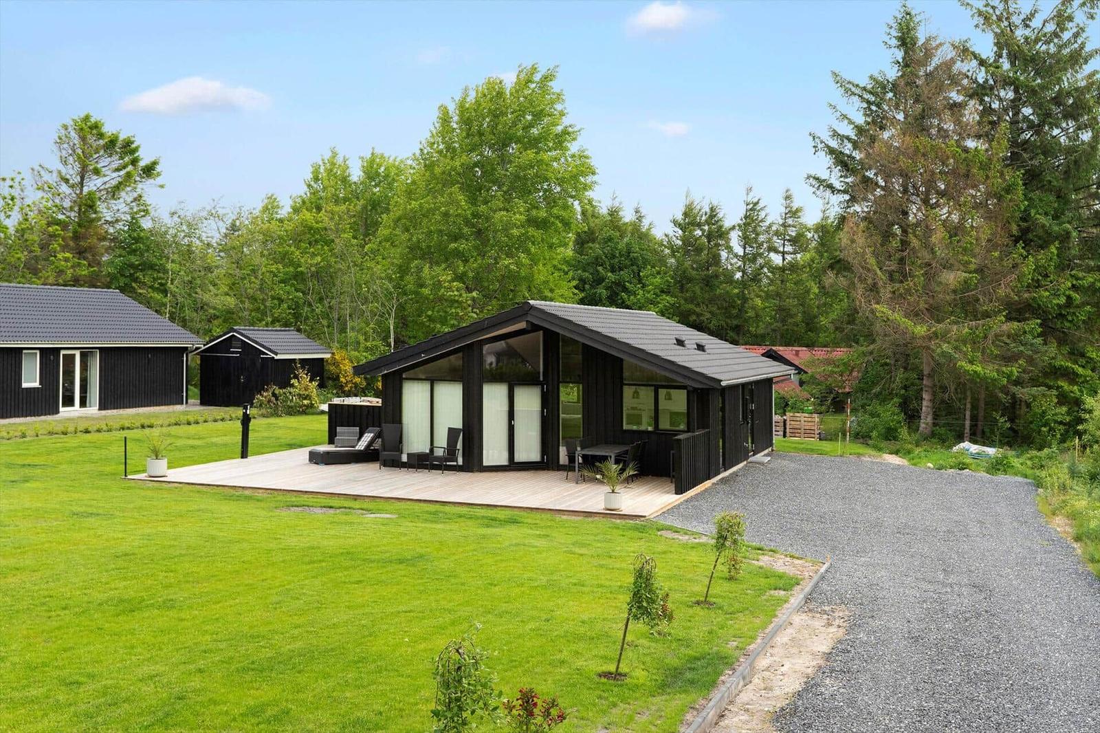 Modern black wooden cabin with large balcony and garden. Surrounded by trees and grass.