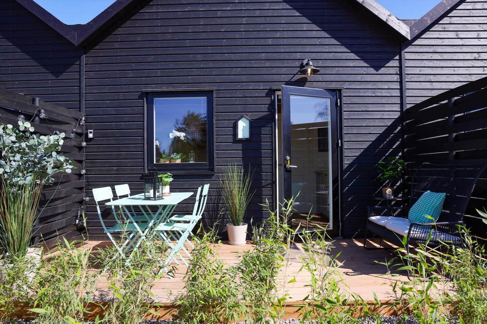 Black wooden cabin with terrace, table, chairs, and seating. Plants and garden area.