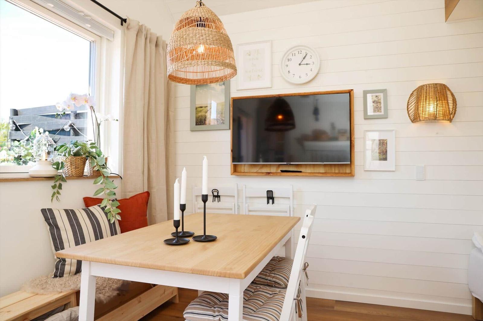 Dining room with wooden table, chairs, and wall-mounted TV. Window with plants and wall lamps.