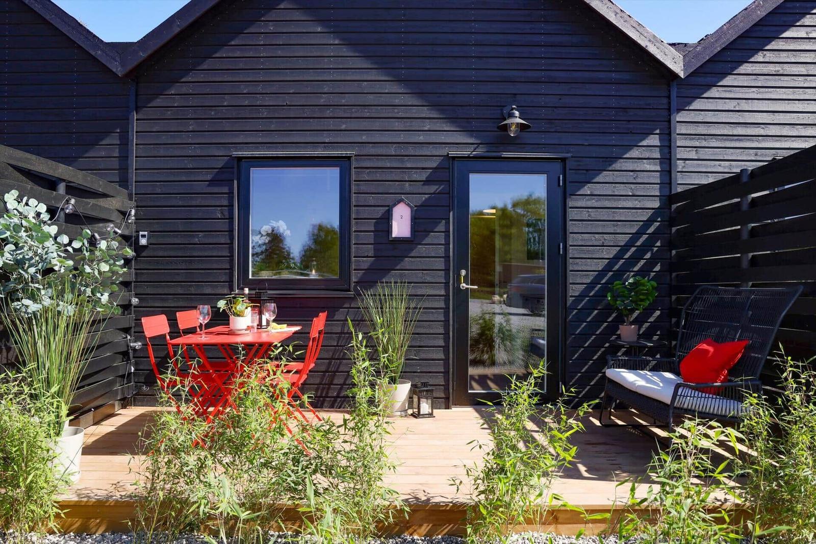 Black wooden house with terrace, red table and chairs, seating area and plants.
