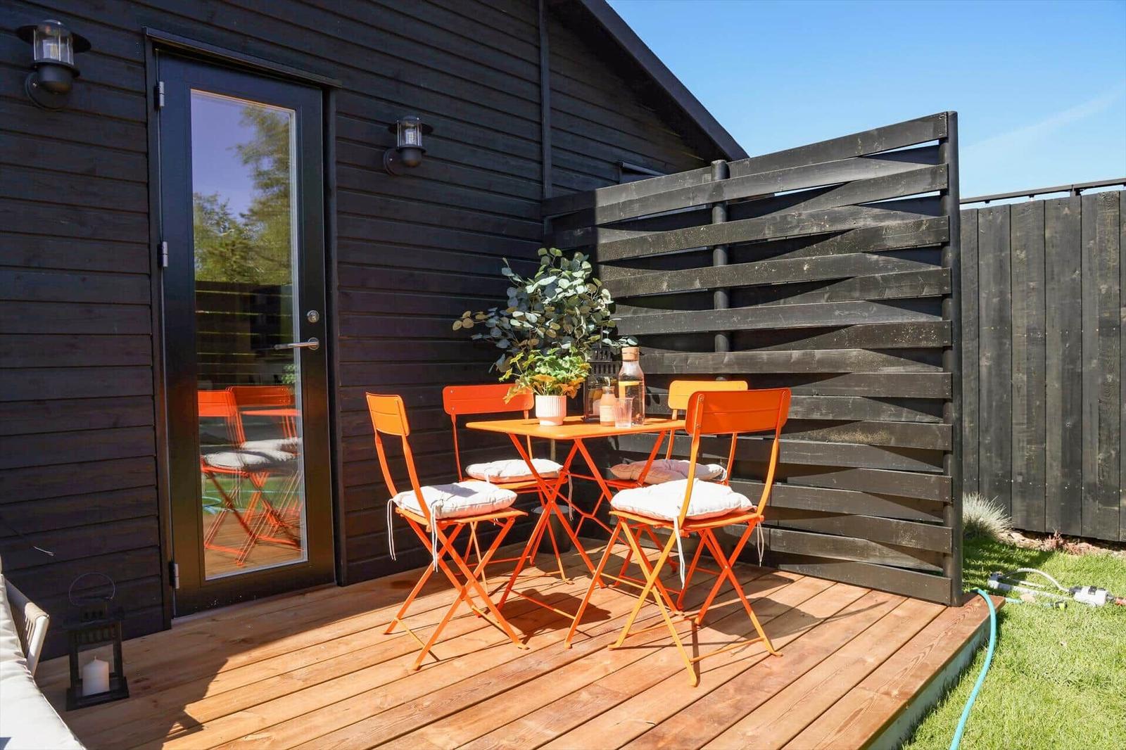 Terrace with orange table and chairs, plant and table decor. Wooden wall and glass door.