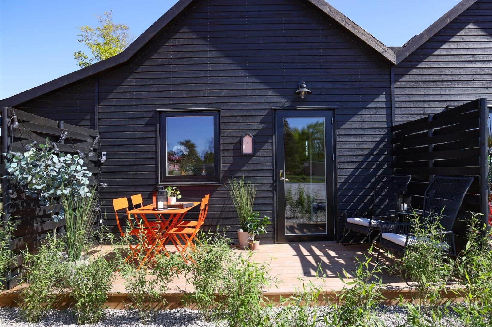 Black wooden house with terrace, orange table and chairs, seating areas, and plants.