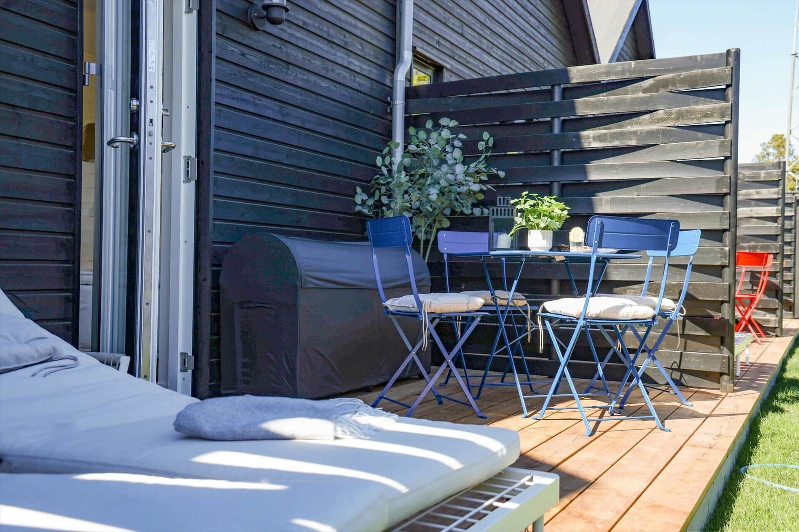 Deck with blue table and chairs, sun lounger, and plants in front of dark wood house.