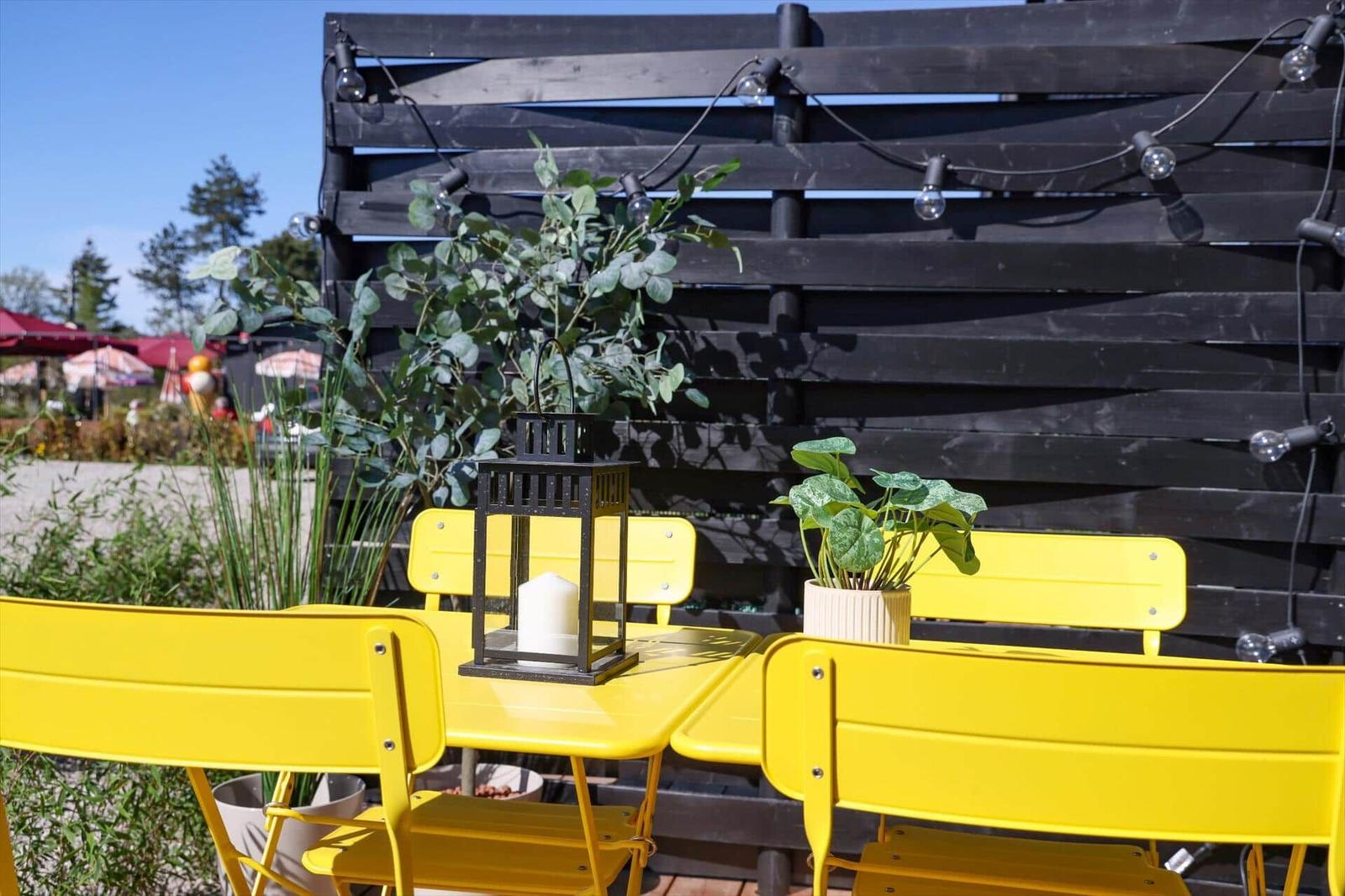 Yellow garden area with table, chairs, plants, and lantern in front of black wooden fence.