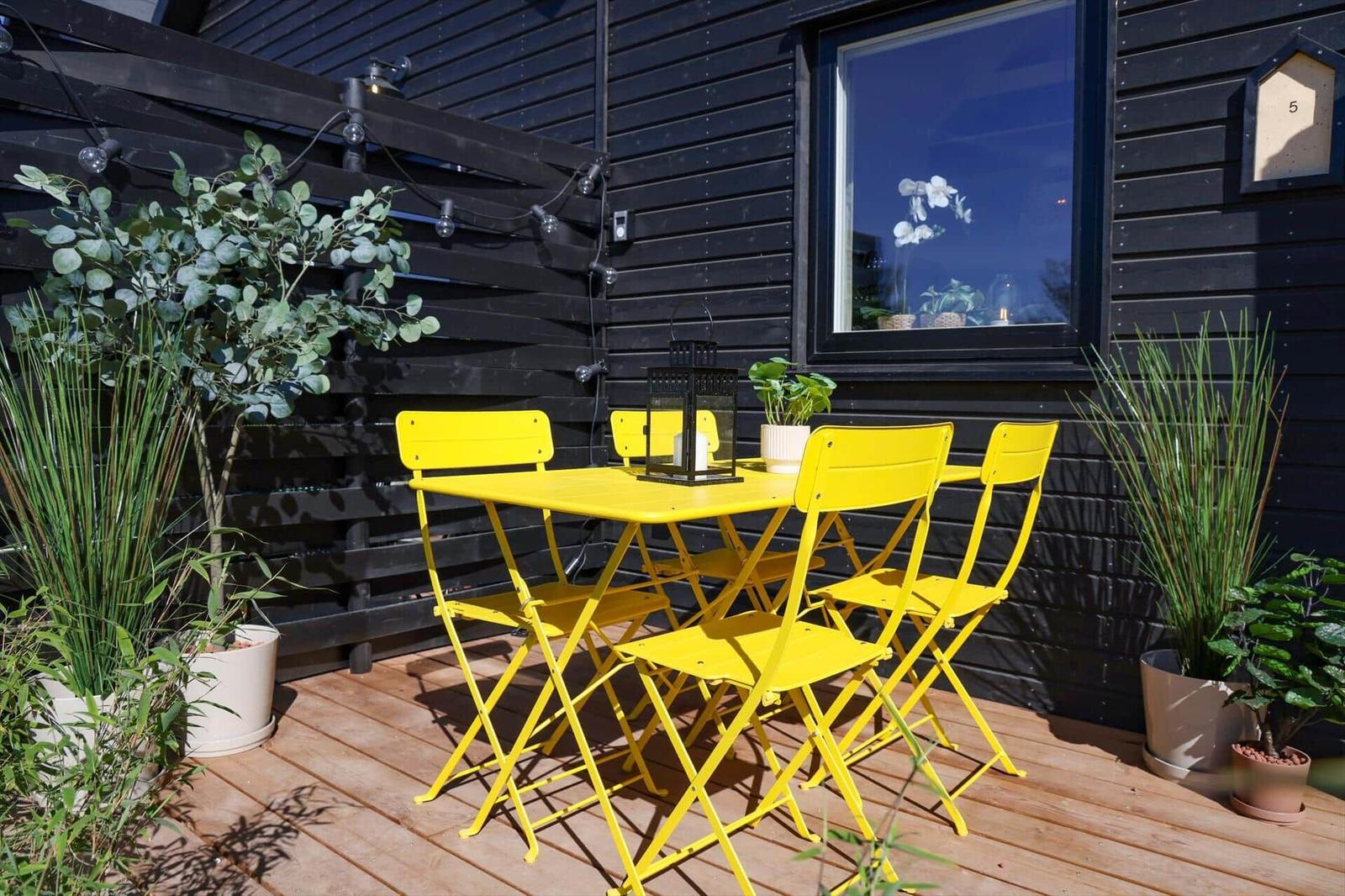 Outdoor area with yellow table and chairs, plants, and window on black wall.