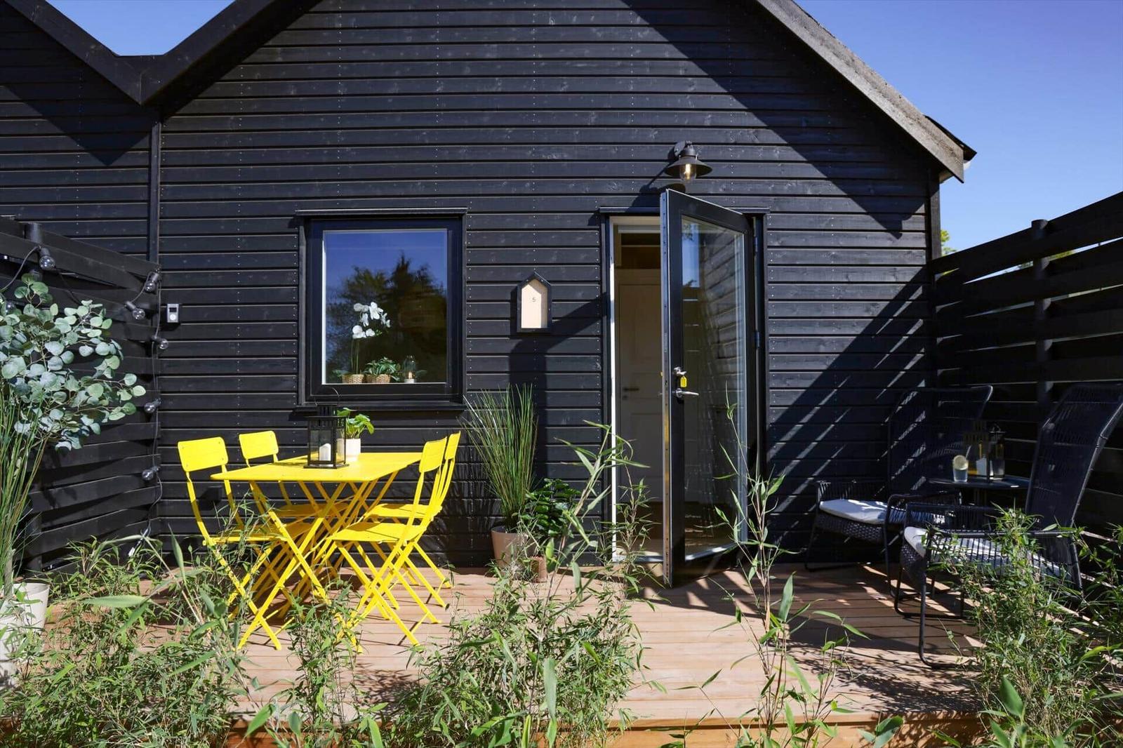 Black wooden house with terrace, yellow table and chairs, plants, and open entrance.