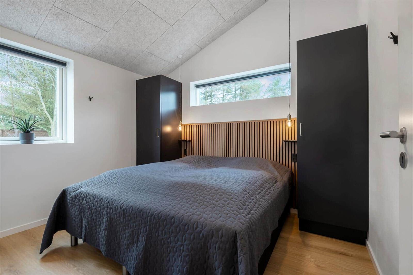 Schlafzimmer mit Bett, zwei Fenstern und zwei schwarzen Schränken.