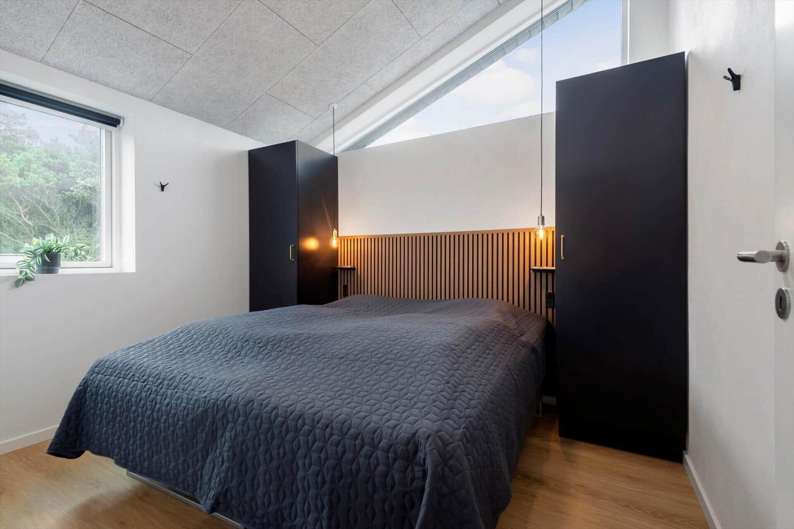 Schlafzimmer mit Doppelbett, Holzfußboden und großen Fenstern. Zwei schwarze Schränke und Holzpaneelen an der Wand.