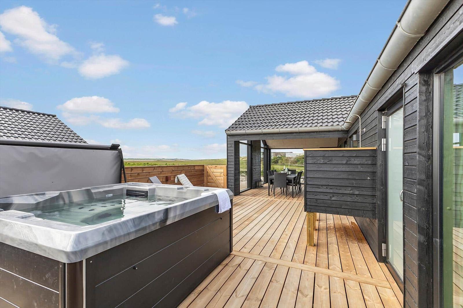 Terrasse mit Whirlpool und Sitzbereich. Holzdecke und schwarze Fassade. Blick auf grüne Landschaft.