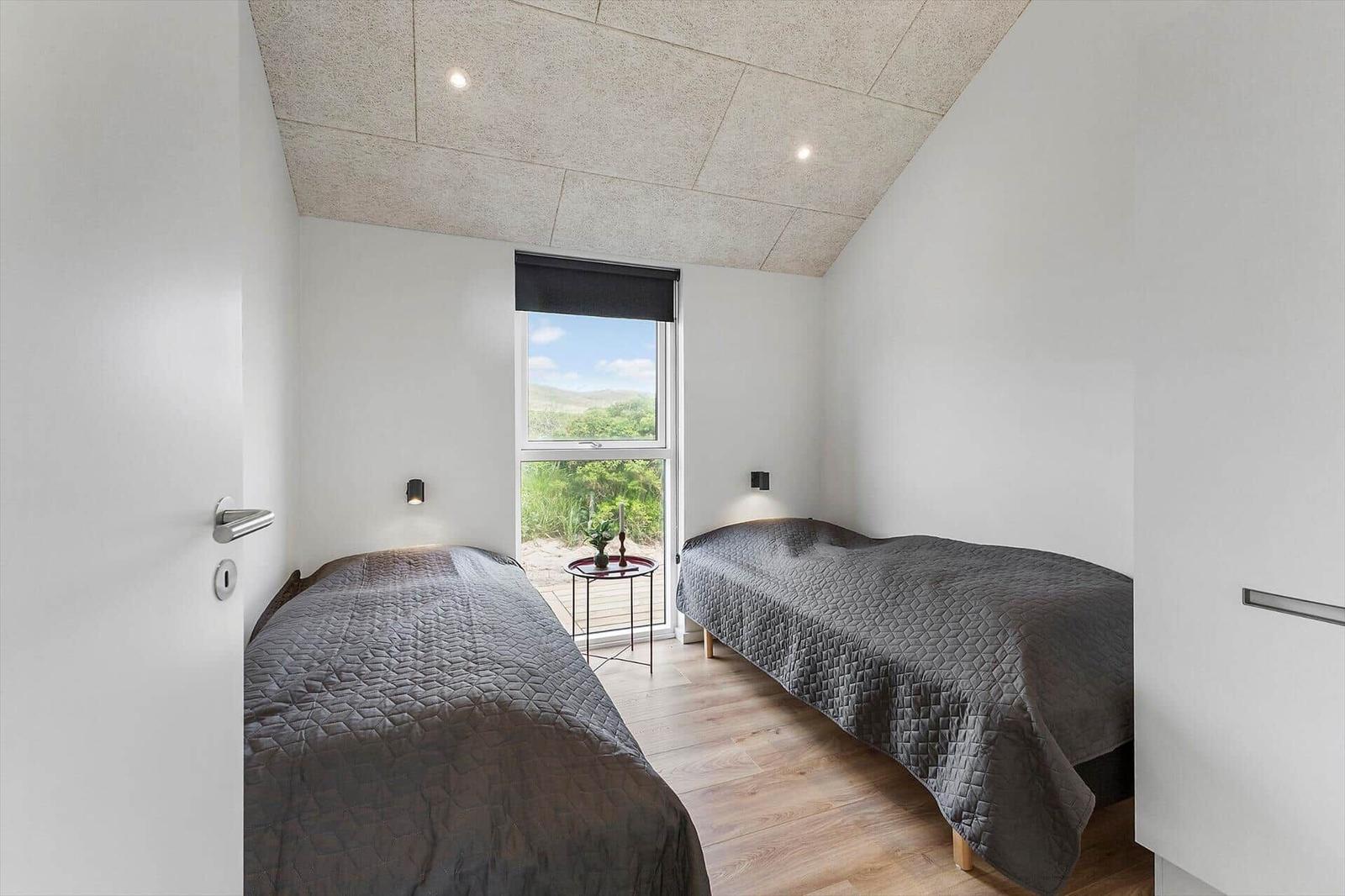 Zwei Betten mit grauer Bettwäsche, Fenster mit Blick auf Landschaft, Holzboden und Deckenleuchten.