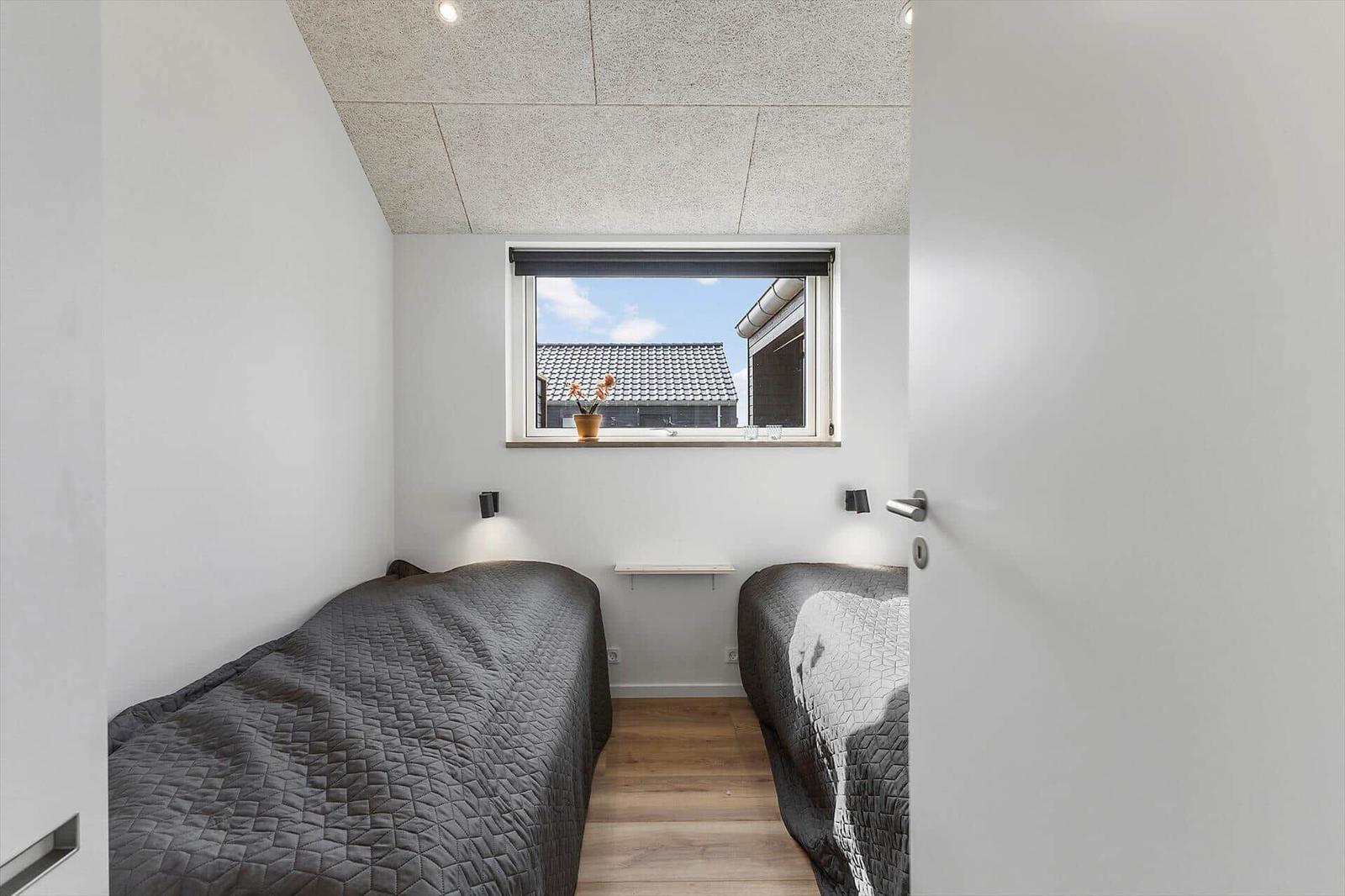 Zwei Betten mit grauen Bettdecken, Fenster mit Blick auf Dächer und Pflanze auf dem Fensterbrett.