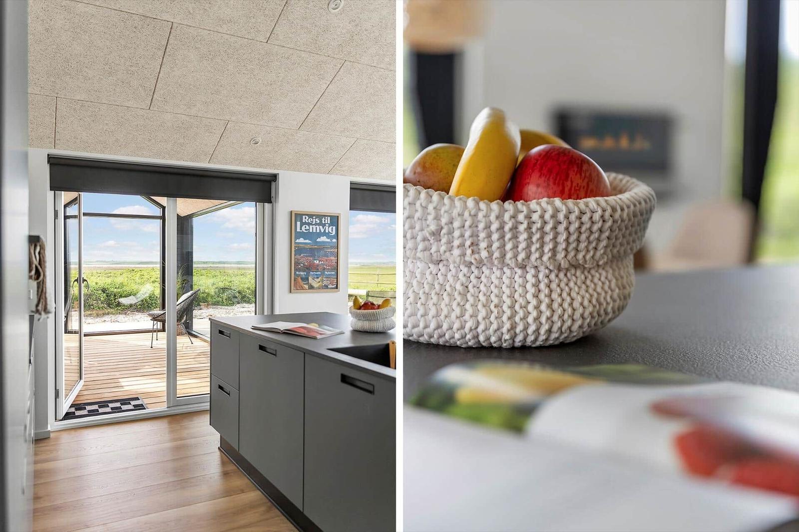 Küche mit Blick auf Terrasse und Landschaft. Obstschale auf Tisch. Holzboden und moderner Küchenbereich.