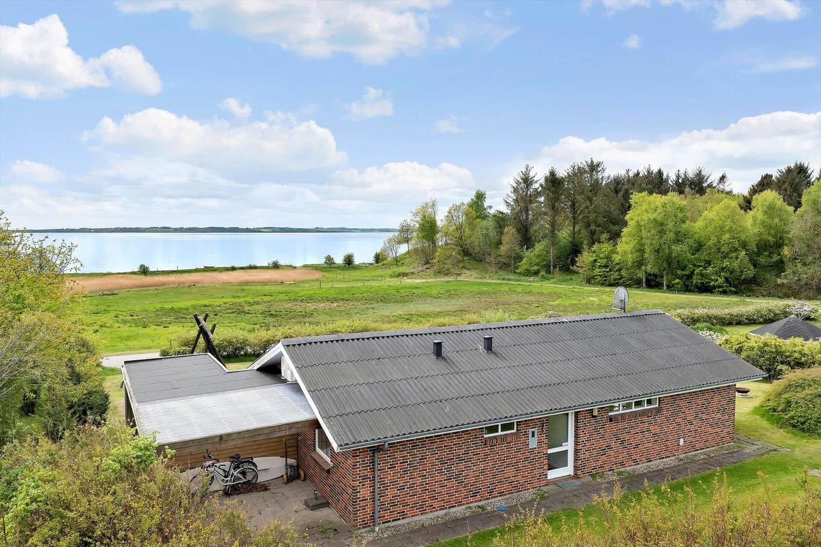 A red brick building with a gray roof, surrounded by green fields and a lake.