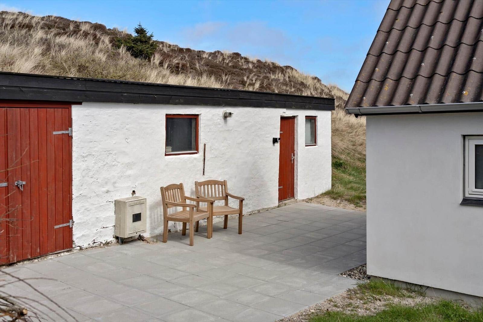 A white house with a red door and two wooden chairs on a stone patio.