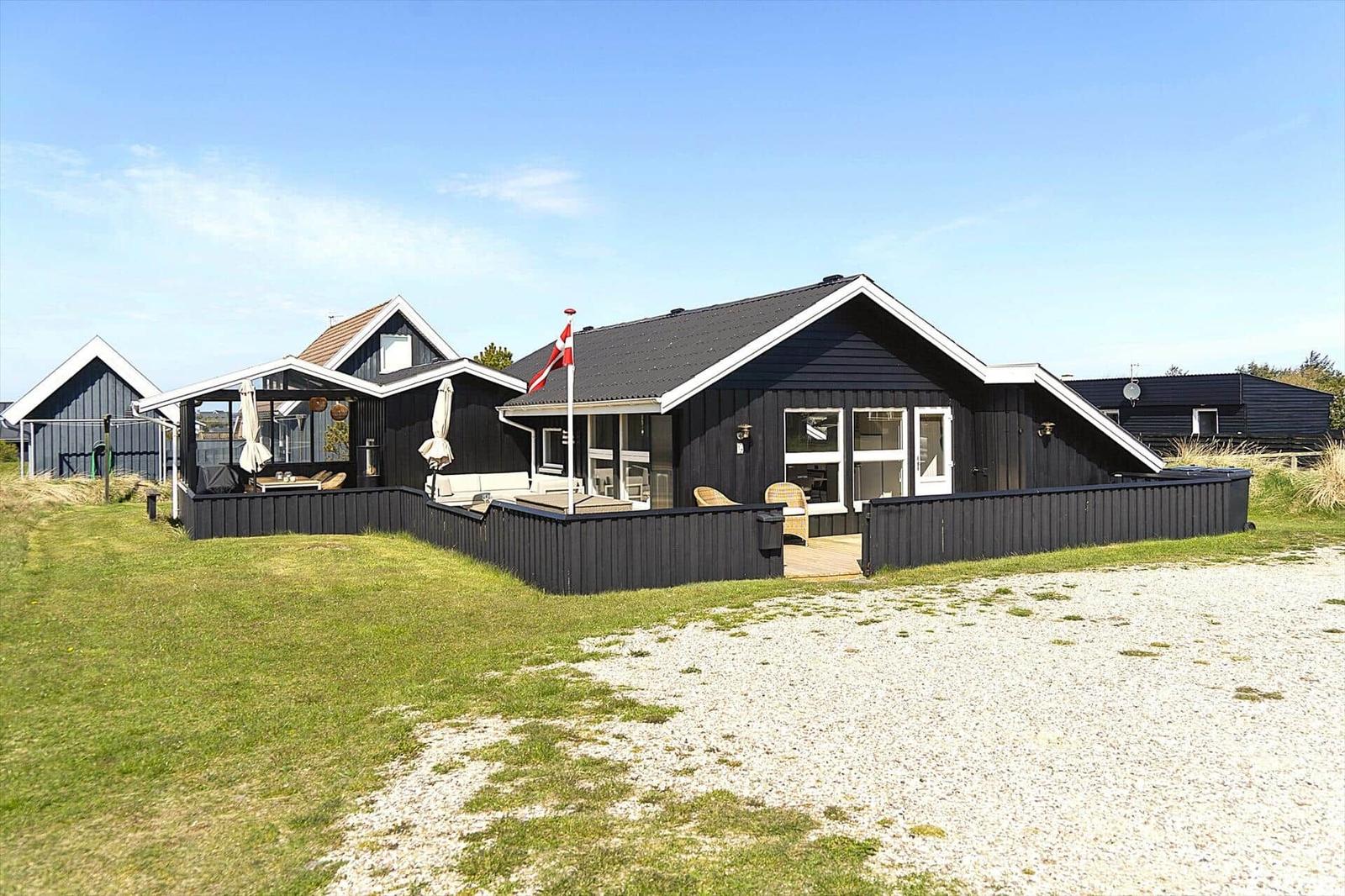 Schwarze Holzhütte mit Terrasse und Dachterrasse. Dänische Flagge weht. Gras und Kies vor dem Haus.