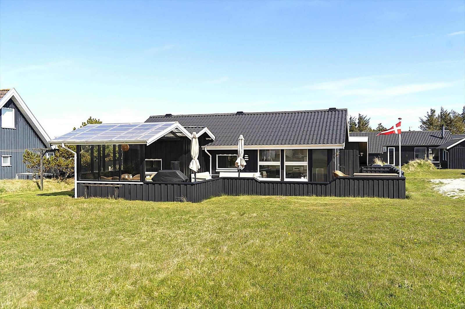 Schwarzes Ferienhaus mit Solaranlage und Terrasse. Grasfläche und dänische Flagge.